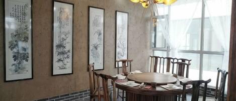 A dining area with a round table surrounded by chairs, framed artwork on the wall, and a large window with sheer curtains.