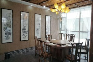 A dining area with a round table surrounded by chairs, framed artwork on the wall, and a large window with sheer curtains. - Shenghua Culture International Hotel (Linze)