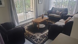 A living room with a sofa set, a coffee table, and a patterned rug. There is a sliding glass door leading to an outdoor area. - Fruitful paradise 
Charming house with WiFi 
welcoming you to enjoy and relax (Church Corner)