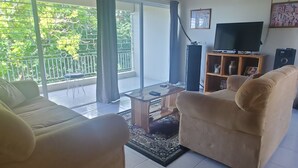 A living room with a sofa, a coffee table, and a television. There is a balcony with a view of greenery. - Fruitful paradise 
Charming house with WiFi 
welcoming you to enjoy and relax (Church Corner)