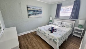 A bedroom with a bed, two bedside tables, a nightstand, a window with curtains, and a painting on the wall. - Parrot Cottage - Margaritaville District (Fort Myers Beach)