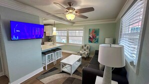 A modern living room with a flat-screen TV, a ceiling fan, a dining area with a table and chairs, and a kitchen with a window. - Parrot Cottage - Margaritaville District (Fort Myers Beach)
