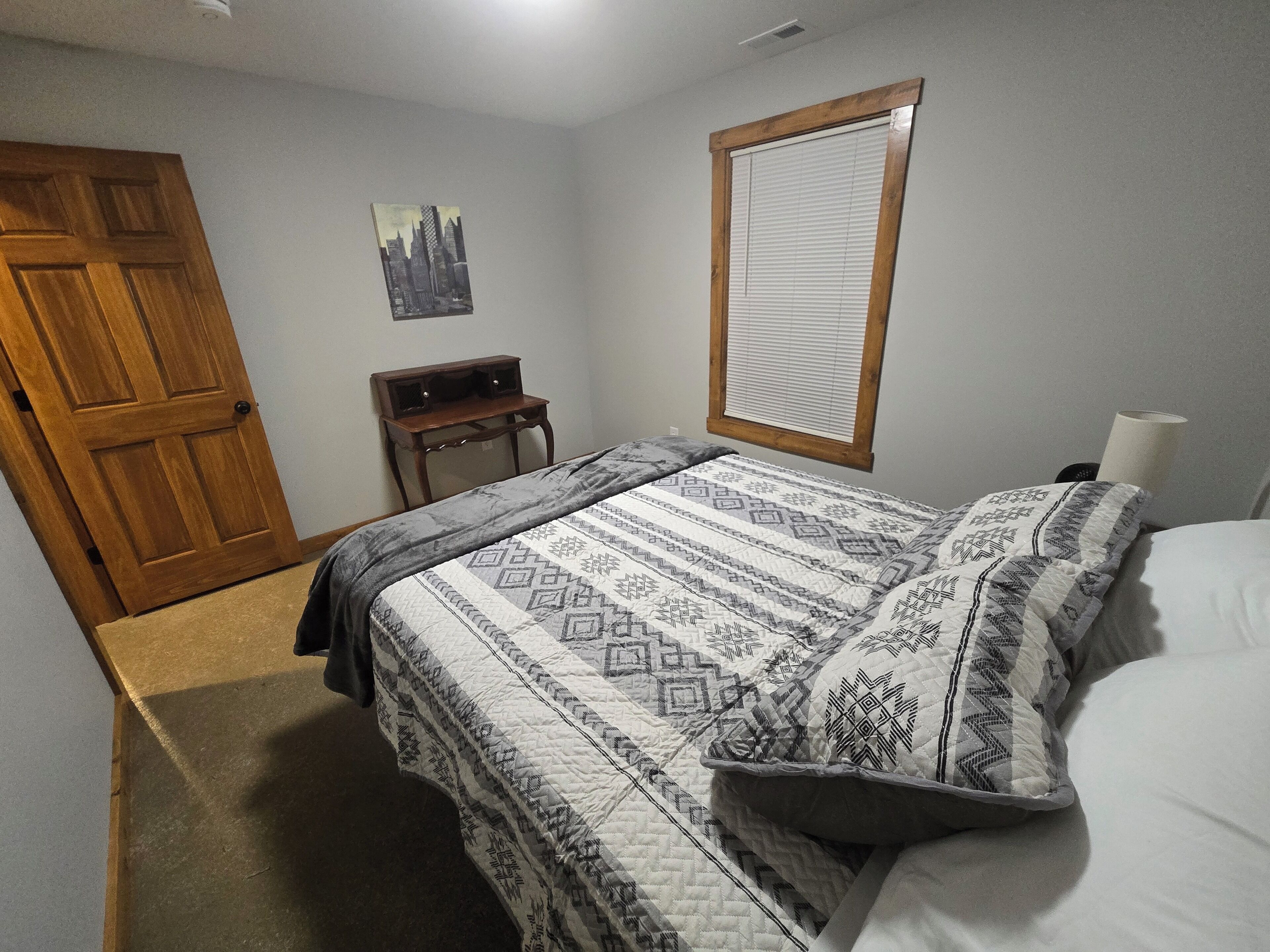 Un dormitorio con cama, una puerta de madera, una mesita, un estante en la pared y una ventana con persianas.