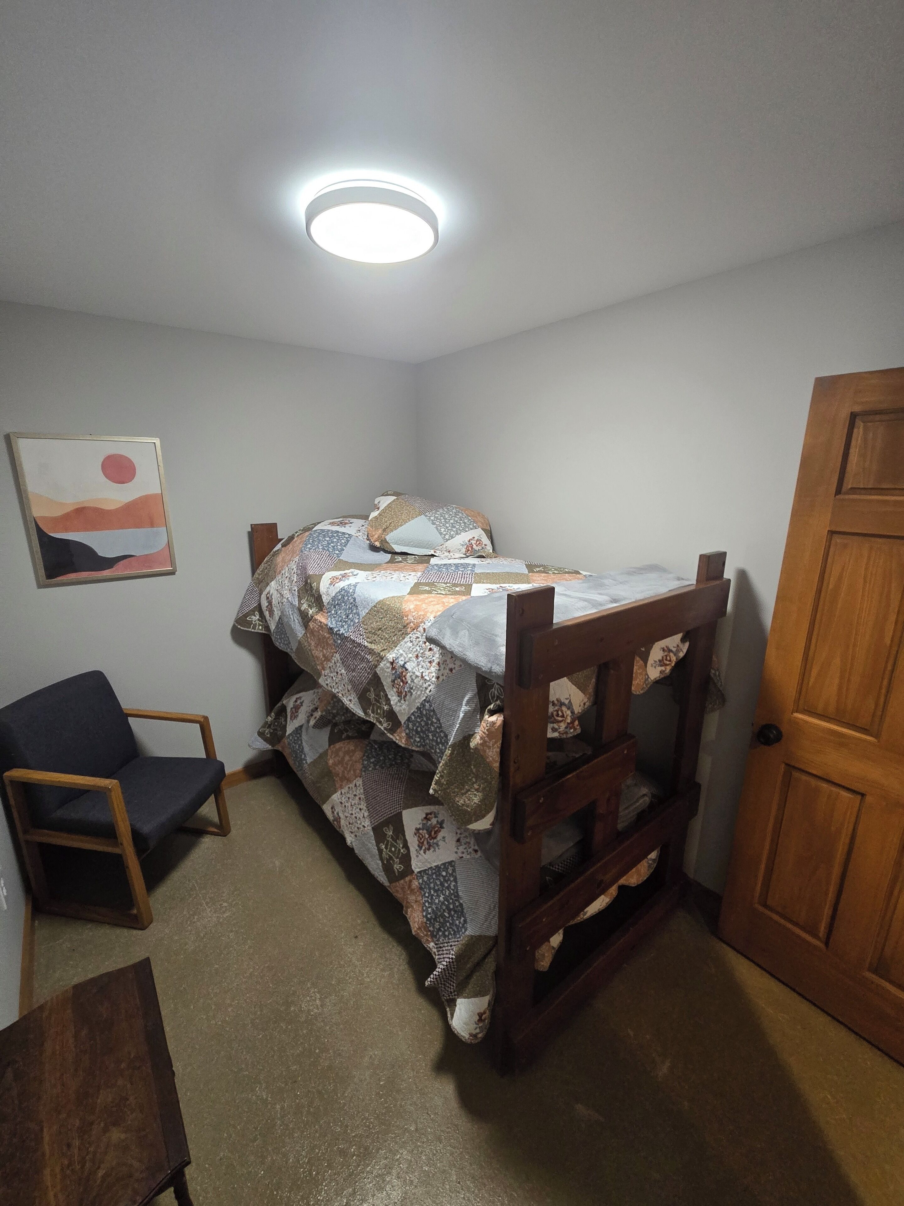 Um quarto pequeno e com pouca luz, com uma cama de solteiro, uma cadeira de madeira, uma porta de madeira e um quadro na parede.