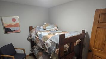 Um quarto pequeno e com pouca luz, com uma cama de solteiro, uma cadeira de madeira, uma porta de madeira e um quadro na parede.