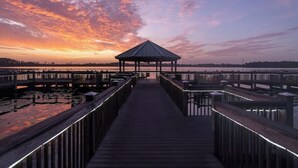 Eine Holzstege führt zu einem Gazebo mit einem Metalldach, umgeben von Wasser und einem Sonnenuntergangshimmel.