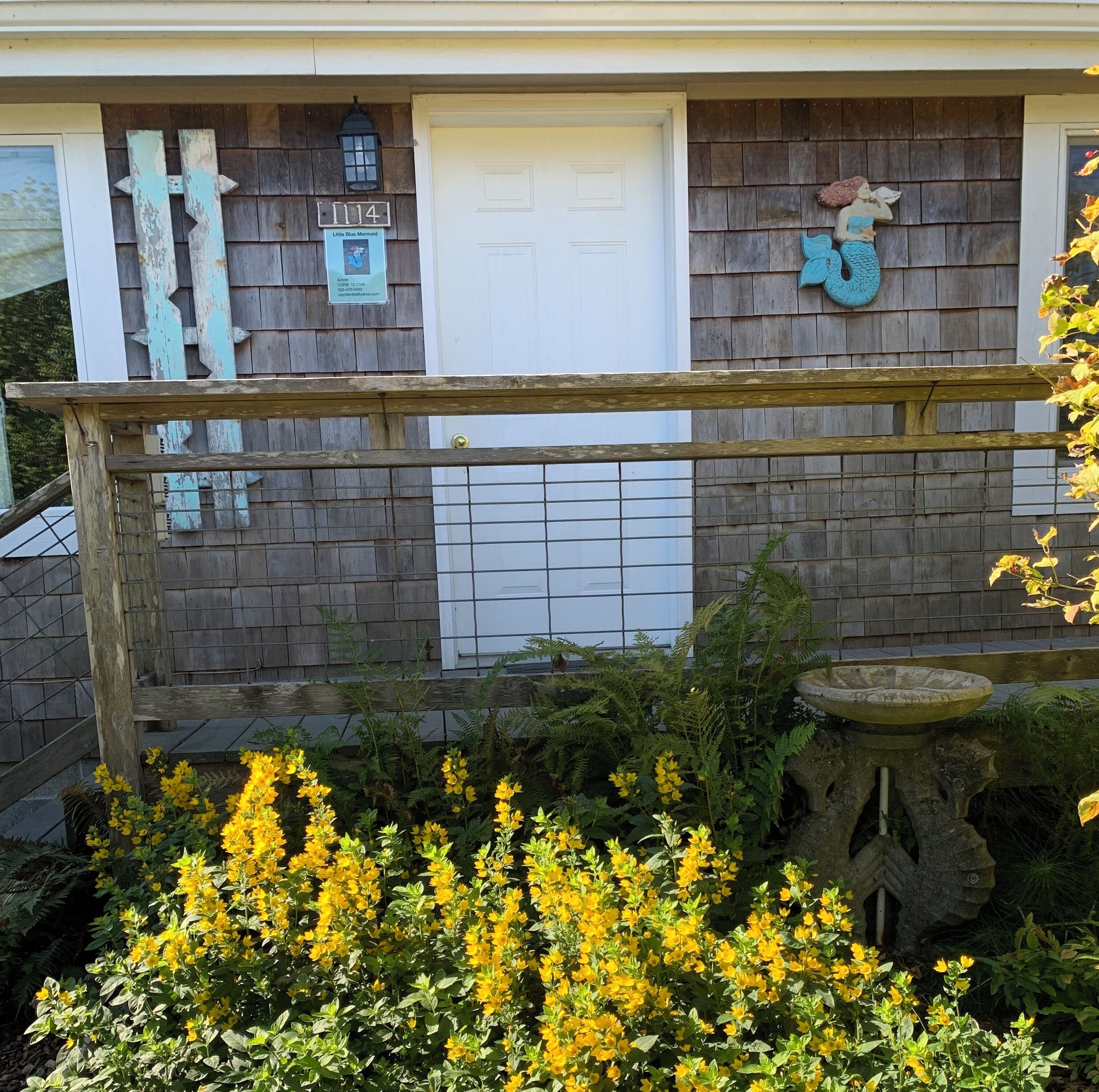 Uma casa com fachada de madeira, uma porta branca e uma caixa de correio. Há uma cerca de madeira em frente à casa.