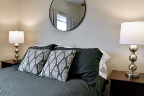 A neatly made bed with a dark green bedspread and patterned pillows, flanked by two bedside tables with lamps. A round mirror hangs on the wall above the bed.