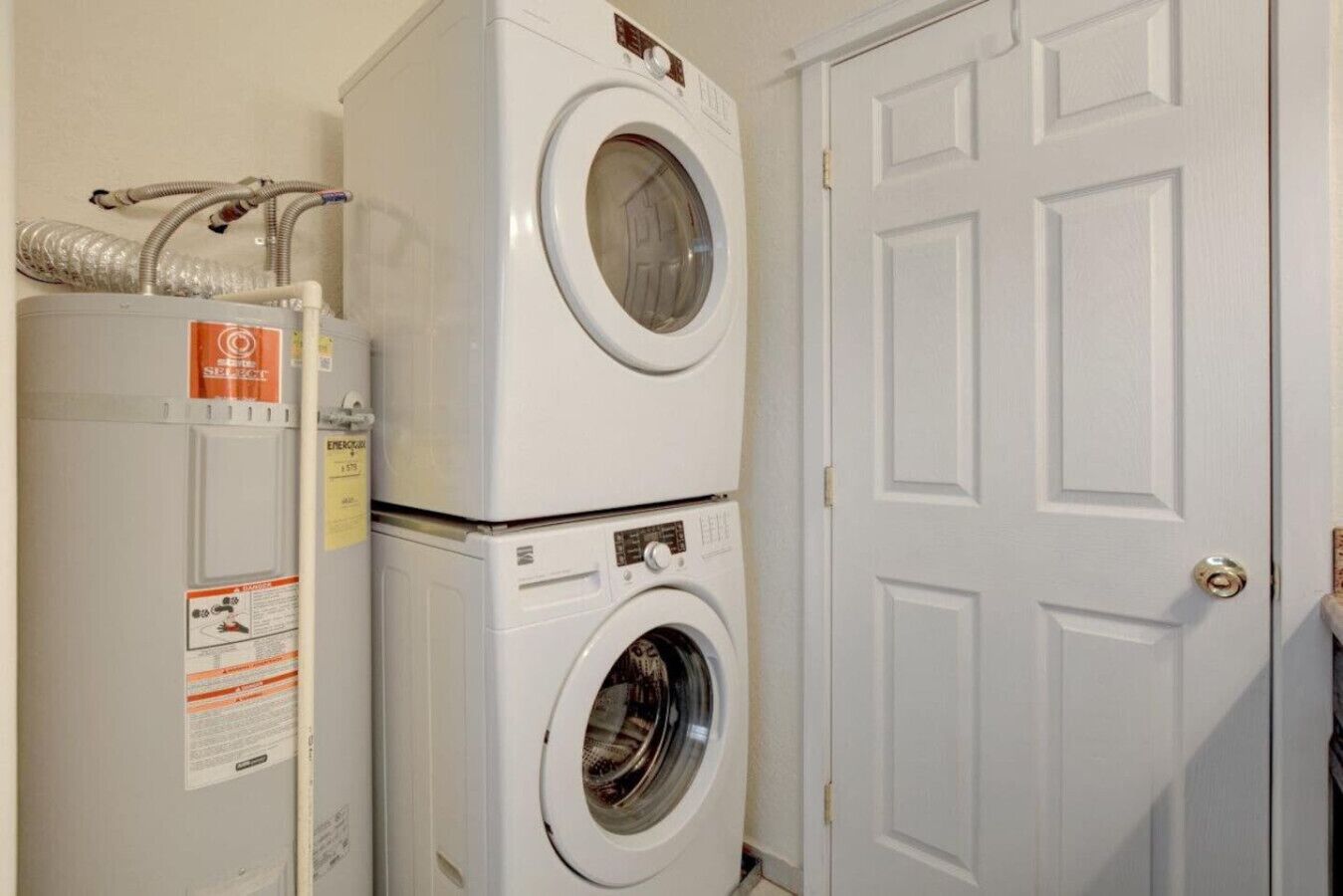 Un área de lavandería con electrodomésticos apilados, incluyendo una secadora y una lavadora, junto a una puerta blanca.