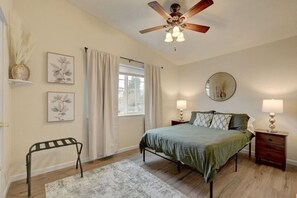 Ein Schlafzimmer mit einem Bett, Nachttischen, einem Deckenventilator, einem Fenster mit Vorhängen und Wandbildern.