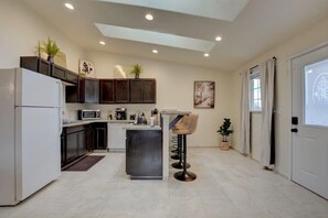 A modern kitchen with dark wooden cabinets, a white refrigerator, a microwave, and a coffee machine. There is a small island with a sink and a chair. The room has a tiled floor and a ceiling with recessed lighting. - Beautiful Loft Apartment w/Views in Medford, OR! (Medford)