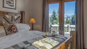 Quarto com uma cama, mesa de cabeceira com abajur, quadro na parede e janela com vista para montanhas cobertas de neve.