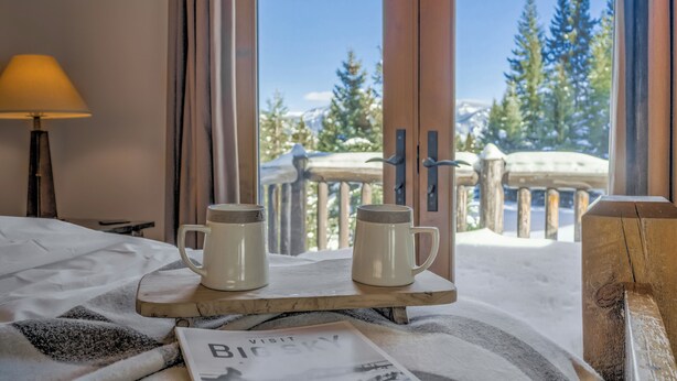 Uma cama com uma bandeja de madeira contendo duas canecas, um abajur e um jornal com a palavra "VISIT" visível.