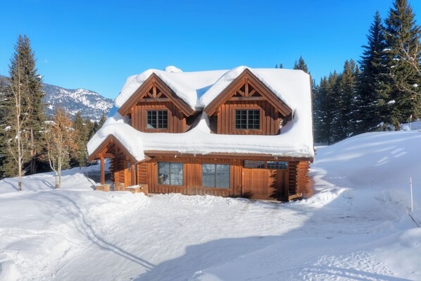 A wooden cabin with a snow-covered roof and large windows, surrounded by snow and trees. - Charming 3-bedroom log cabin with Hot Tub and fitness room in delightful Big Sky (Big Sky)