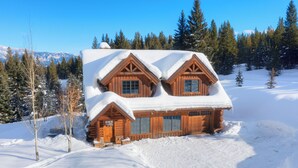 Uma cabana de tronco com design simétrico, janelas grandes e telhado coberto de neve, rodeada por uma paisagem nevada e árvores perenes.