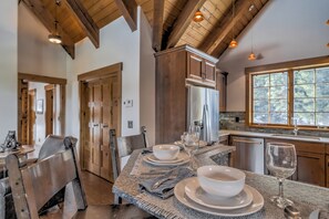 A dining area with a table set for two, wooden chairs, and a view of the kitchen and dining room through a window. - Charming 3-bedroom log cabin with Hot Tub and fitness room in delightful Big Sky (Big Sky)
