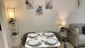 Un comedor con una mesa preparada para cuatro personas, rodeada de sillas grises, un sofá y una pequeña mesita con una lámpara.
