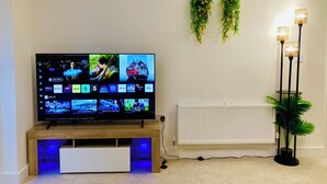 Un televisor de pantalla plana sobre un soporte de madera con retroiluminación azul, un radiador blanco y una lámpara de pie con tres luces.