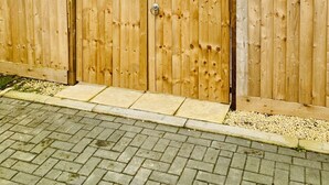 Una valla de madera con un portón, un área pavimentada con grava, y un edificio al fondo.