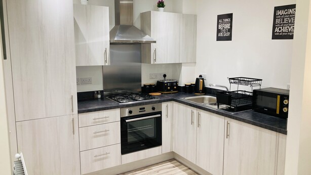 Una cocina moderna con armarios blancos, una campana extractora de acero inoxidable, una estufa a gas, un horno empotrado y un microondas. Hay dos electrodomésticos negros en la encimera y un cartel blanco y negro en la pared.
