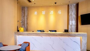 Una zona de recepción de hotel con mostrador de mármol, un televisor montado en la pared y un área de descanso con sillas naranjas y una mesa redonda.