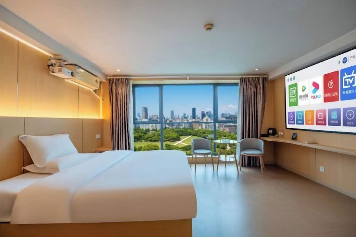 Hotelkamer met een groot bed, een bureau met stoel, een televisiescherm waarop verschillende apps worden weergegeven, en een stadszicht door grote ramen.