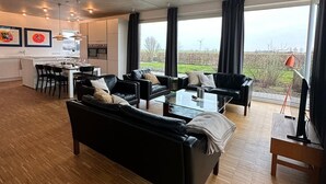 A spacious living area with a large black sofa, a dining table with chairs, and a kitchen in the background.