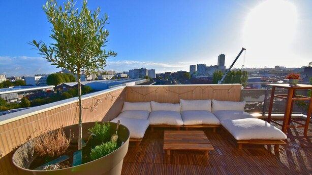 Una terrazza sul tetto con un pavimento in legno, un grande vaso con un albero, una zona salotto con cuscineria bianca e vista sullo skyline della città.