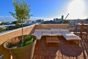 A rooftop terrace with a wooden deck, a large planter with a tree, a white cushioned seating area, and a view of the city skyline. - Le Belvédère Parisien - Billiards and Large Terrace (Aubervilliers)