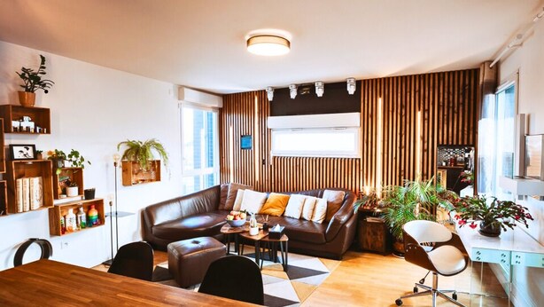 A modern living room with a brown leather sofa, a wooden dining table, and a white desk. There are multiple plants and a large window providing natural light. - L'Échappée Parisienne - Canal St-Denis (Aubervilliers)