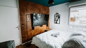 A bedroom with a wooden panel wall, a large flat-screen TV, a bed with floral bedding, and a window with a view of buildings. - L'Échappée Parisienne - Canal St-Denis (Aubervilliers)