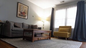 Ett vardagsrum med en grå soffa, ett soffbord i trä, en gul fåtölj och ett stort fönster med gardiner.