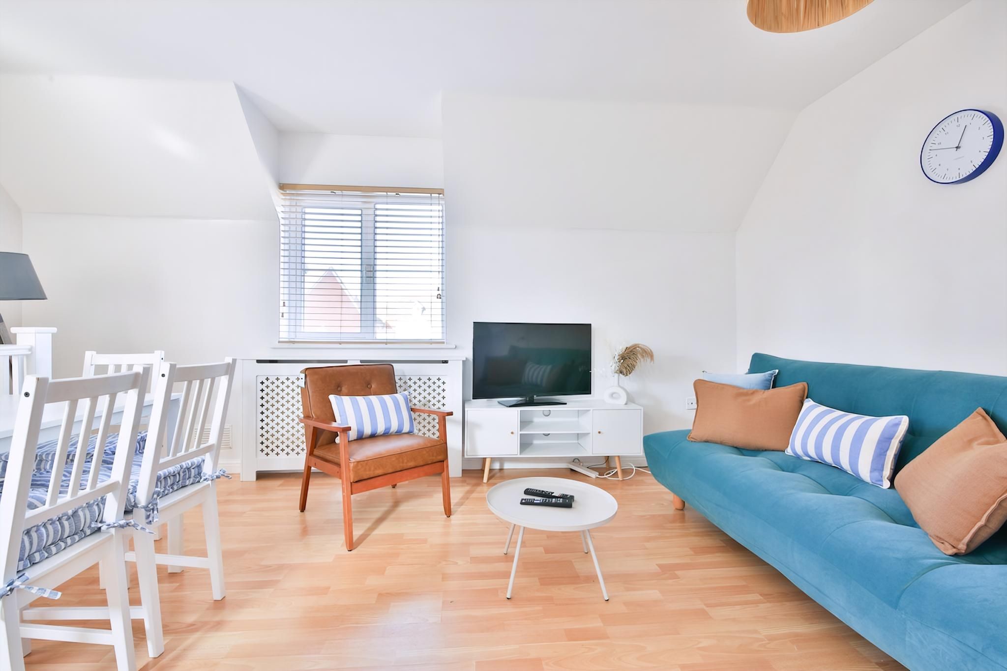 A modern living room with a blue sofa, white chairs, a TV, and a clock on the wall.