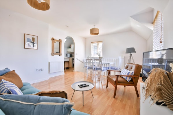A modern living room with a sofa, a coffee table, a dining area with chairs, a TV, and a kitchen in the background. - 2 Bed House Near Beach with Free Parking (Barry)