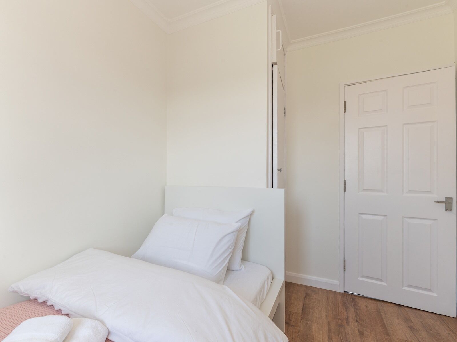 A bedroom with a single bed, white bedding, a wooden floor, and a white door.