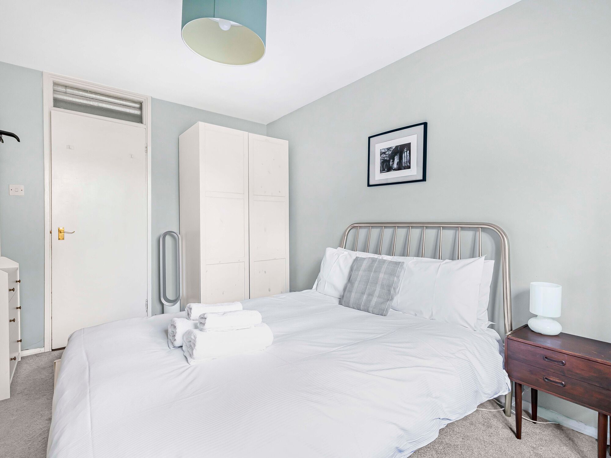 A neatly made bed with white linens and a metal headboard, a wooden nightstand with a lamp, a white wardrobe, and a framed picture on the wall.