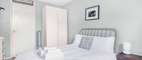 A neatly made bed with white linens and a metal headboard, a wooden nightstand with a lamp, a white wardrobe, and a framed picture on the wall.