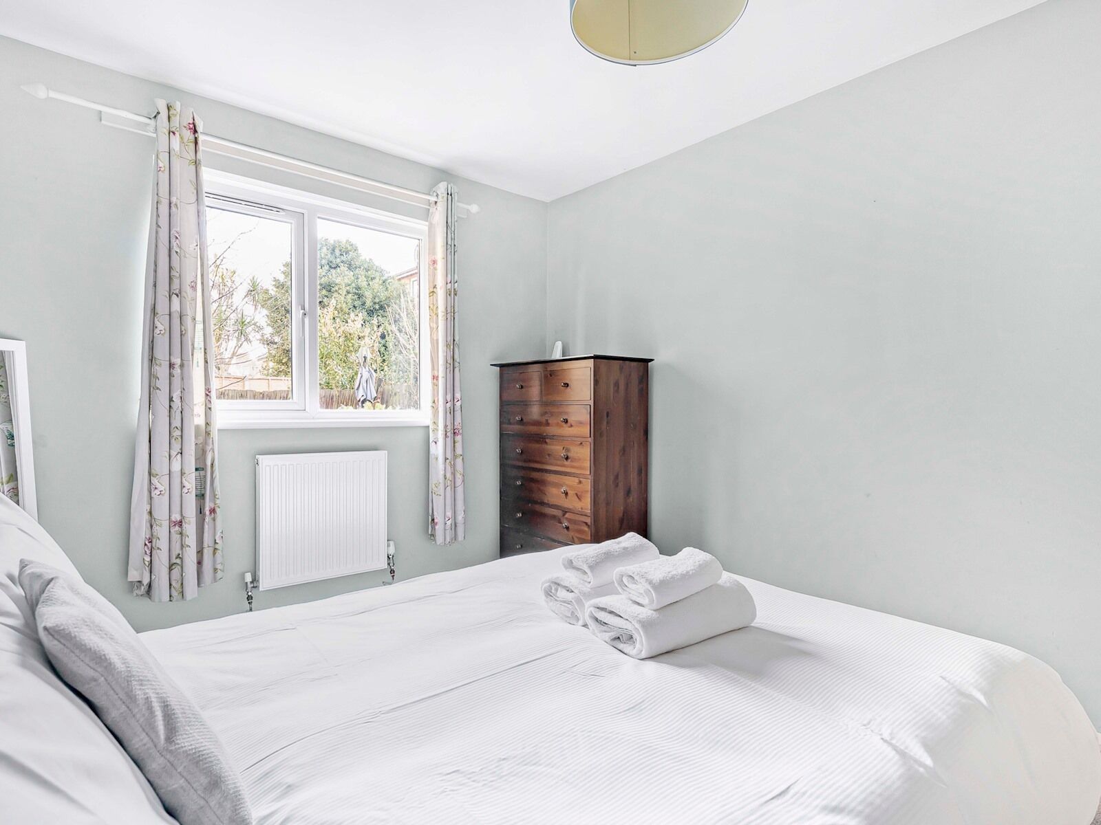 A bedroom with a bed, a dresser, a window with curtains, and a radiator.