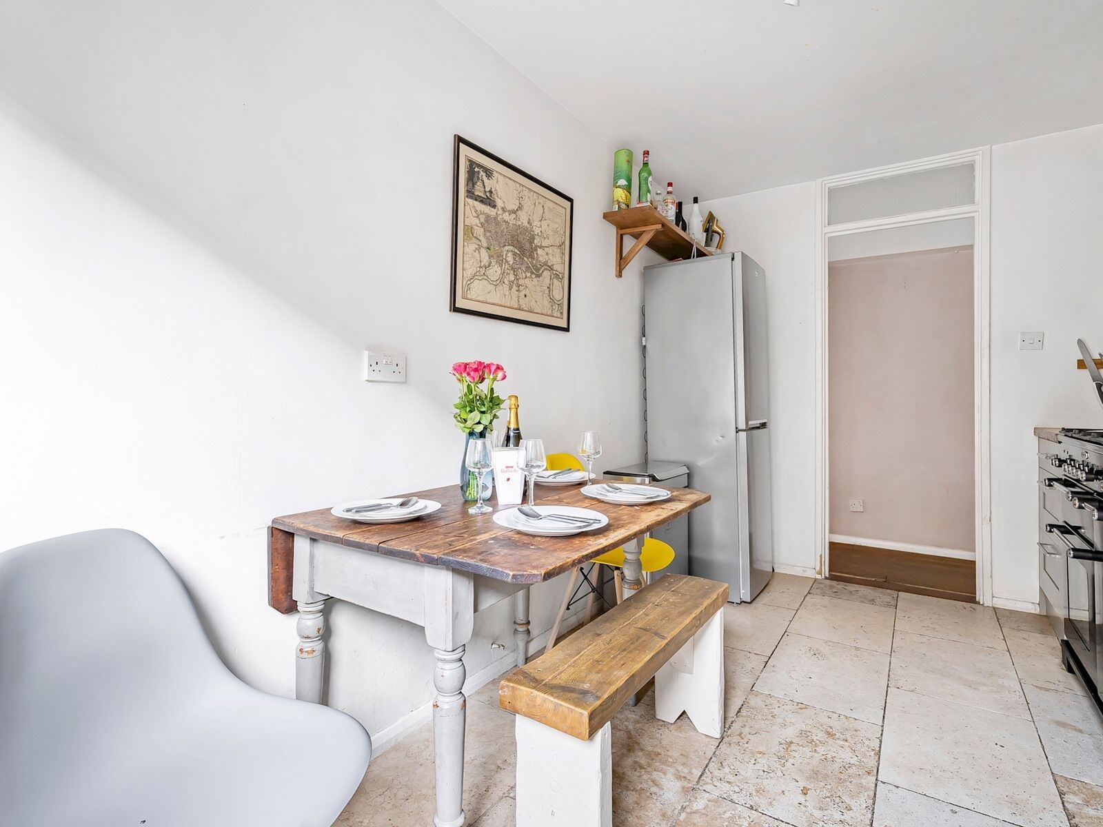 A compact kitchen with a wooden table set for dining, a bench, a refrigerator, and a shelf with bottles.
