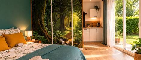 A bedroom with a large bed, a view of a forest through a sliding door, a small kitchenette, and a sliding glass door to the outside.