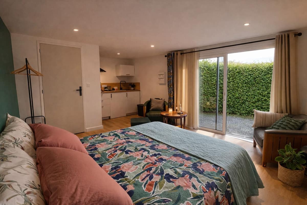 A bedroom with a large bed, a small table, a chair, and a view of a garden through a sliding door.