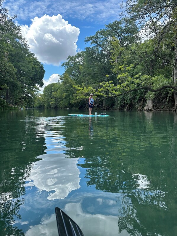 Une personne se tient debout sur une planche de paddle, au milieu d’une rivière paisible, entourée d’une végétation luxuriante.
