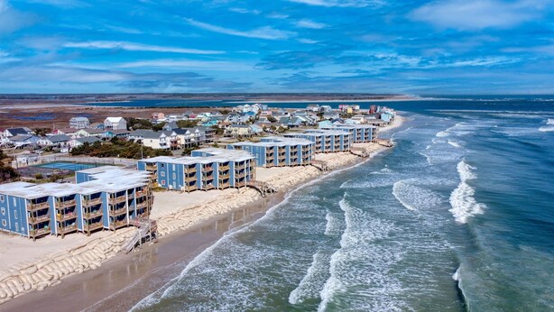 A coastal hotel with multiple blue buildings, a sandy beach, and ocean waves. - Oceanfront Bliss: Sand and Views at Watchin' Waves (North Topsail Beach)