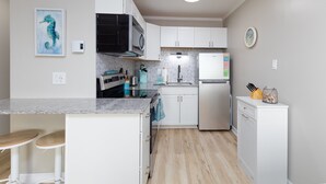 A modern kitchen with white cabinets, a built-in oven, a microwave, and a dishwasher. There is a small island with stools and a painting of a seahorse on the wall. - Oceanfront Bliss: Sand and Views at Watchin' Waves (North Topsail Beach)