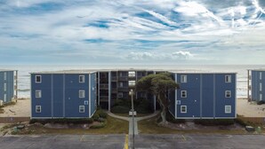 A beachfront apartment complex with multiple floors, a central entrance, and a parking area in front. - Oceanfront Bliss: Sand and Views at Watchin' Waves (North Topsail Beach)
