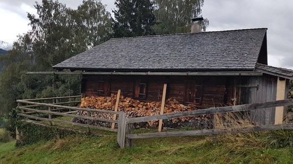 A rustic wooden cabin with a sloped roof, surrounded by a wooden fence and a stack of firewood.