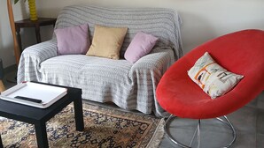 A living room with a grey sofa, a red chair, a patterned rug, and a small table with a remote control. - Small house by the sea Pets welcome (Pornic)