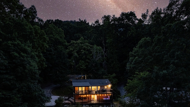 Une cabane à deux étages dotée d’un balcon, entourée d’une forêt dense, sous un ciel étoilé.