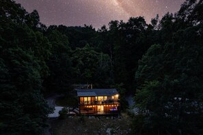 Una cabaña de dos pisos con balcón, rodeada de un denso bosque, bajo un cielo estrellado.