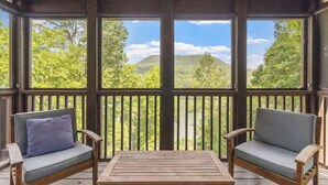 Une terrasse en bois avec deux fauteuils rembourrés et une table, offrant une vue panoramique sur un paysage verdoyant, avec des arbres et des collines.